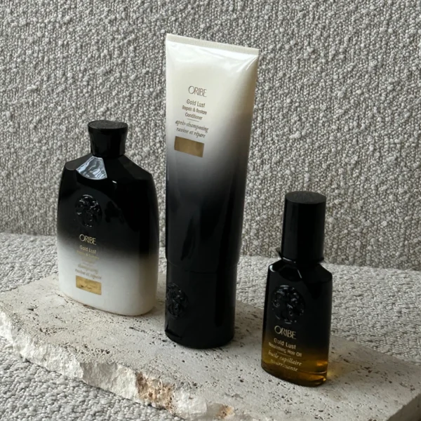 Oribe Gold Lust hårplejeprodukter, herunder shampoo, balsam og hårolie, arrangeret på en stenplade med et struktureret baggrund