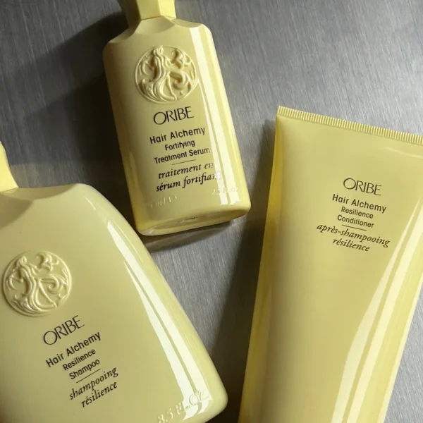 Tre gulde Oribe Hair Alchemy Resilience produkter, herunder shampoo, conditioner og et serum, arrangeret på en metaloverflade